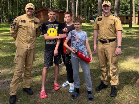 Harcerze i strażacy pozują do wspólnego zdjęcia pamiątkowego w obozowej scenerii.