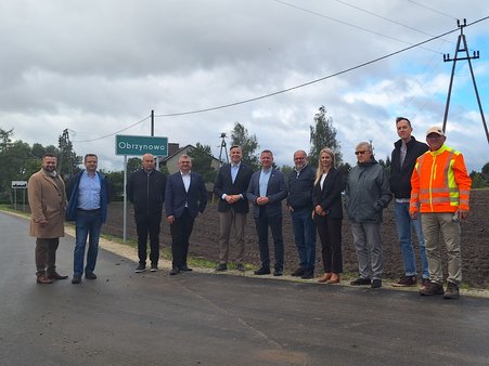 Zdjęcie przedstawia 11 osób (w tym jedna kobieta) stojących na drodze przy tablicy z nazwą miejscowości Obrzynowo i pozujących do zdjęcia, w tle pole i dachy domów