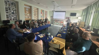 zdjęcie przedstawia grupę osób siedzącą przy stole oraz mężczyznę omawiającego prezentację wyświetloną z rzutnika na ścianie