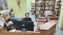 Nauczycielka siedząca przy biurku a w tle regały z książkami wypełniające przestrzeń.