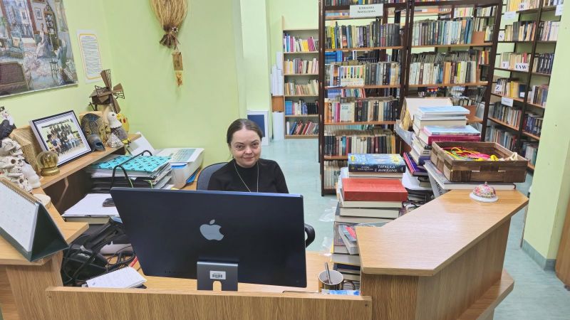 Nauczycielka siedząca przy biurku a w tle regały z książkami wypełniające przestrzeń.
