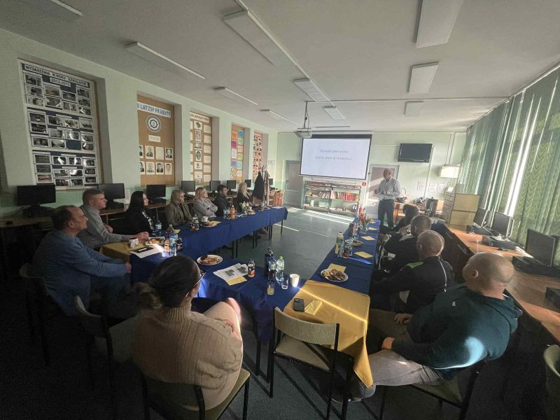 zdjęcie przedstawia grupę osób siedzącą przy stole oraz mężczyznę omawiającego prezentację wyświetloną z rzutnika na ścianie