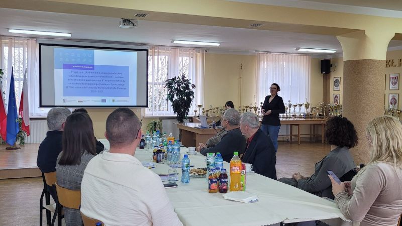 Kobieta prowadzi prezentację o funduszach europejskich przed grupą osób siedzących przy stole w sali z pucharami i flagami Polski oraz Unii Europejskiej.