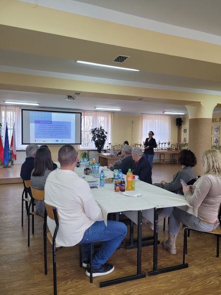 Uczestnicy siedzą przy stole i słuchają prezentacji o projekcie edukacyjnym finansowanym z funduszy europejskich, w jasnej sali z trofeami.