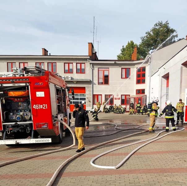 Strażacy w akcji, część przy wozie, jeden trzyma wąż i polewa wodą.