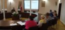 Trzecia grupa szkoleniowa uczestnicząca w szkoleniu na miejscach siedzących przy okrągłym stole konferencyjnym wraz z osobą prowadzącą szkolenie omawiającą szczegółowe tematy szkolenia